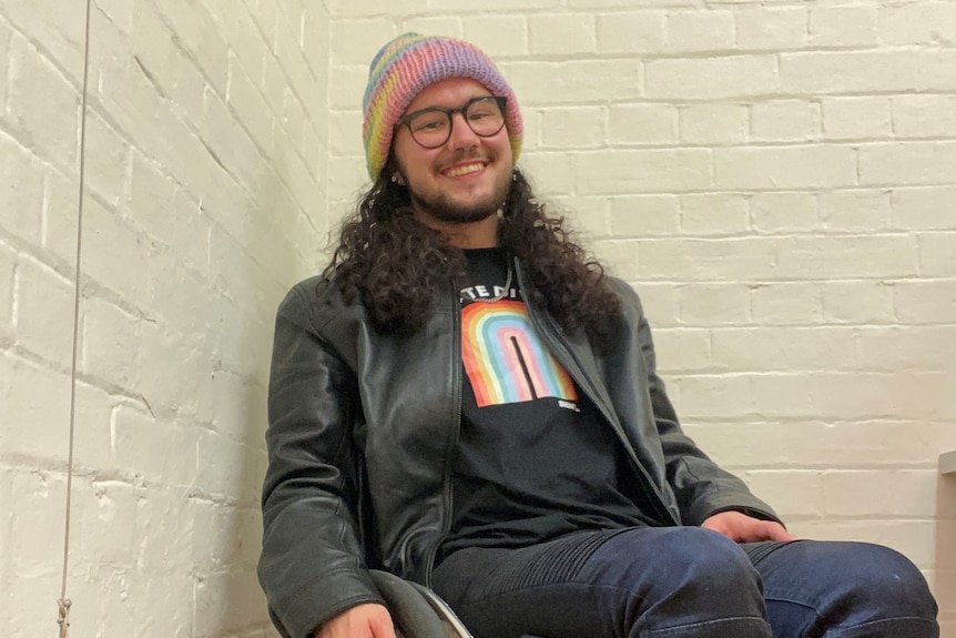 Evan smiling wearing a rainbow tee and beanie while sitting in a wheelchair