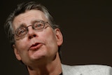 Stephen King wears a suit and glasses on a stage in front of a black background