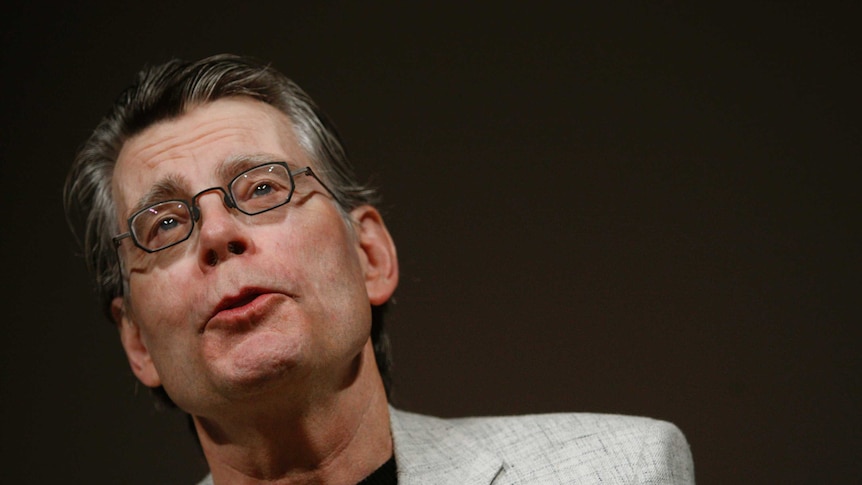 Stephen King wears a suit and glasses on a stage in front of a black background
