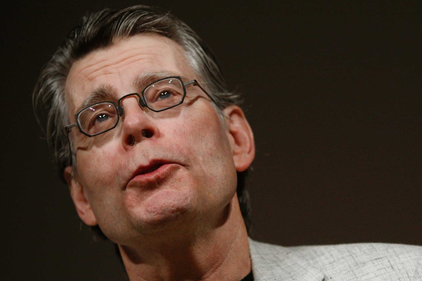 Stephen King wears a suit and glasses on a stage in front of a black background