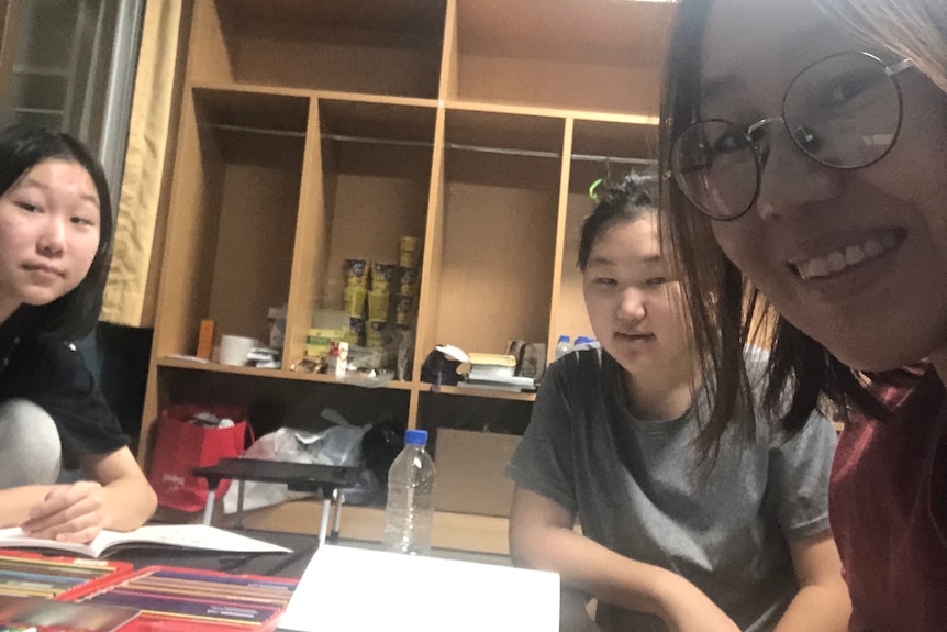 A young woman and two pre-teen girls smile for a selfie at a desk with colouring pencils and paper. Open lockers line the walls