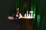 Two ladies wearing feathered and formal attire sit in a dimly lit room.