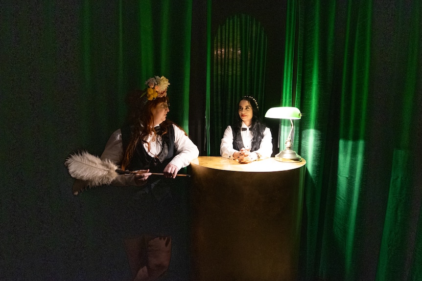 Two ladies wearing feathered and formal attire sit in a dimly lit room.