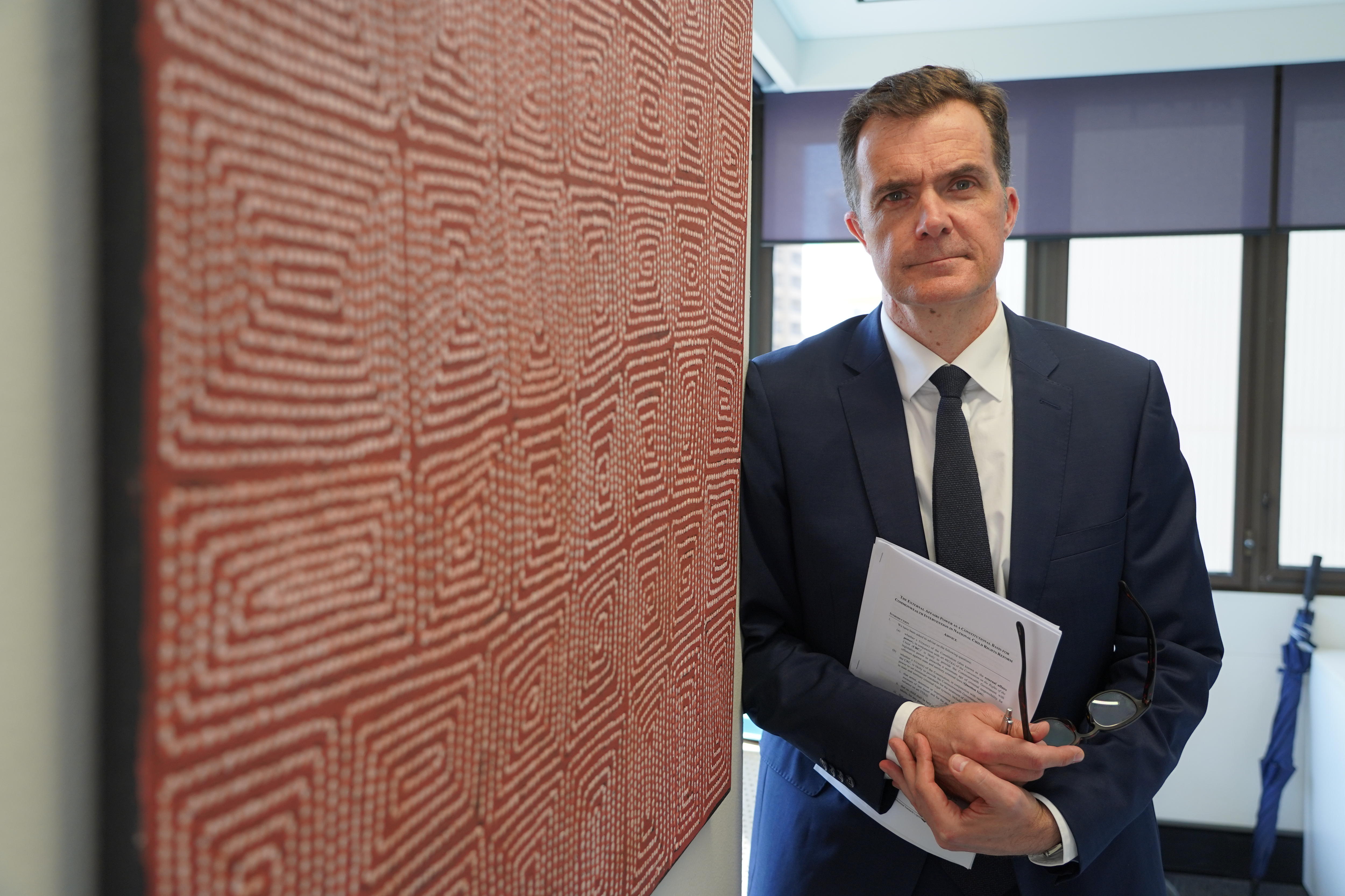 A man in a black suit and tie stands holding papers next to a piece of Aboriginal art