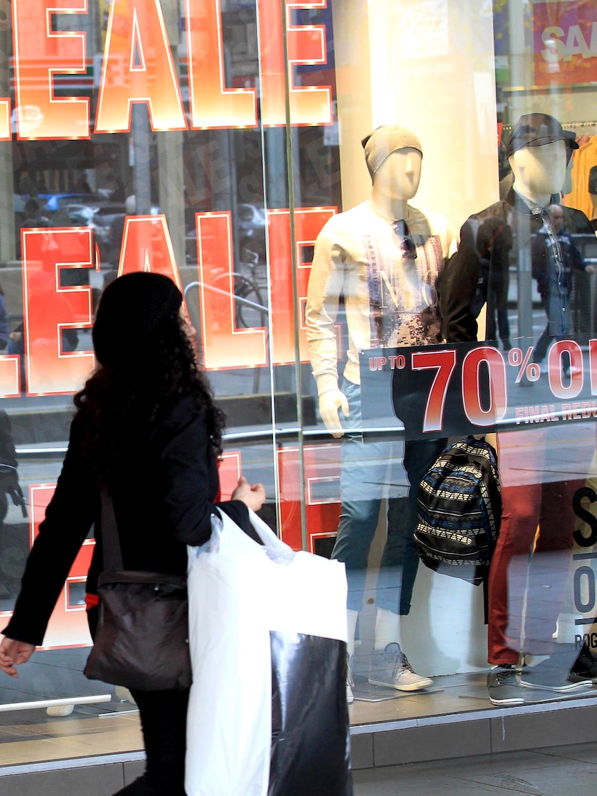 A woman with shopping bags walks past a retail store displaying sale signs.