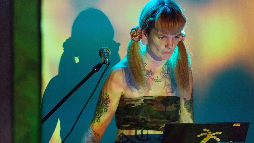 Bridget Chappell, with their hair in pigtails, controls a laptop, which is decorated with stickers. A microphone is to the side