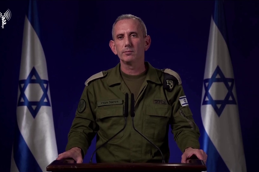 A man in military uniform standing at a podium flanked by two israeli flags