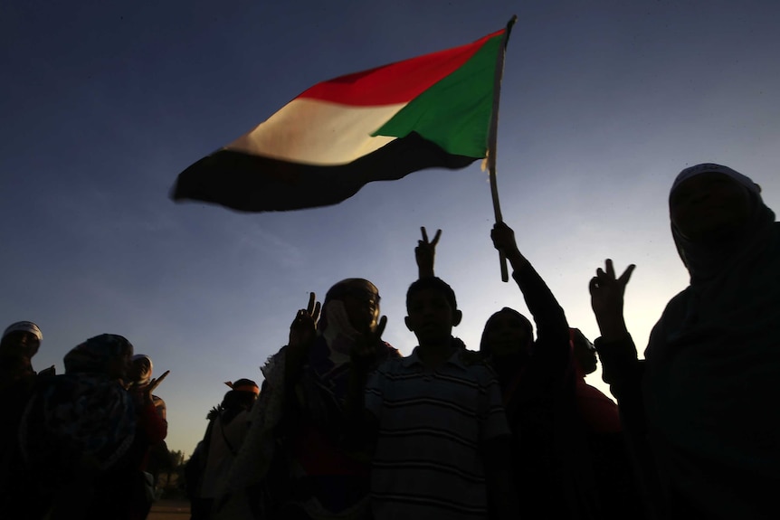 You view a silhouette of a large crowd against a dusk sky with the Sudanese flag flying above them.