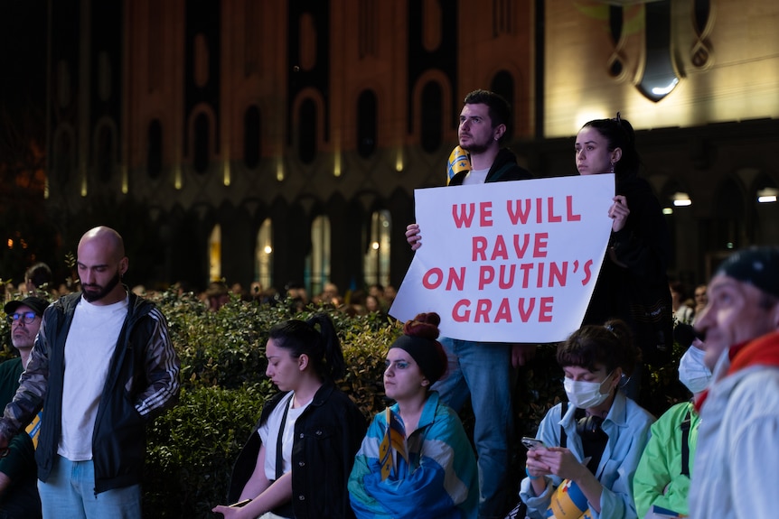 A person holds a poster reading 'we will rave on Putin's grave'.