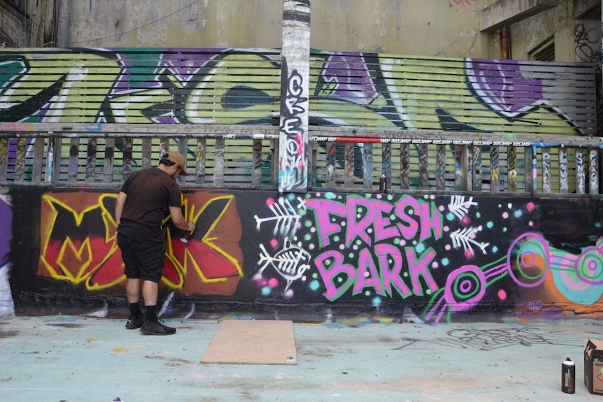 A man spray painting an industrial-looking wall with the words 'Fresh Bark' painted on it.