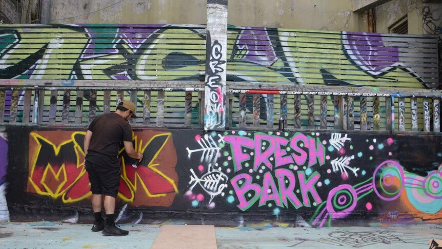 A man spray painting an industrial looking wall with the words 'Fresh Bark' painted on it.
