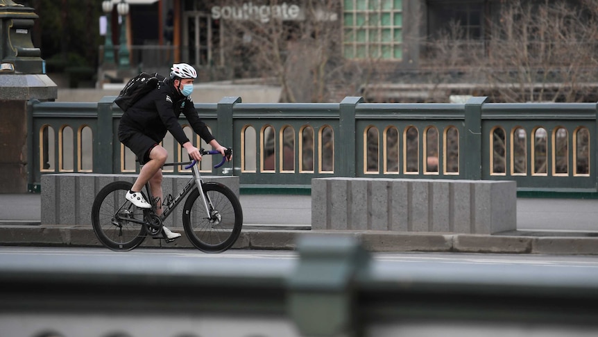 A man rides a bike over a bridge wearing a white bike helmet and a blue surgical face mask. No one else can be seen in the shot