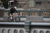 A man rides a bike over a bridge wearing a white bike helmet and a blue surgical face mask. No one else can be seen in the shot