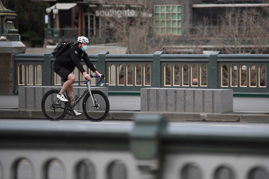 A man rides a bike over a bridge wearing a white bike helmet and a blue surgical face mask. No one else can be seen in the shot