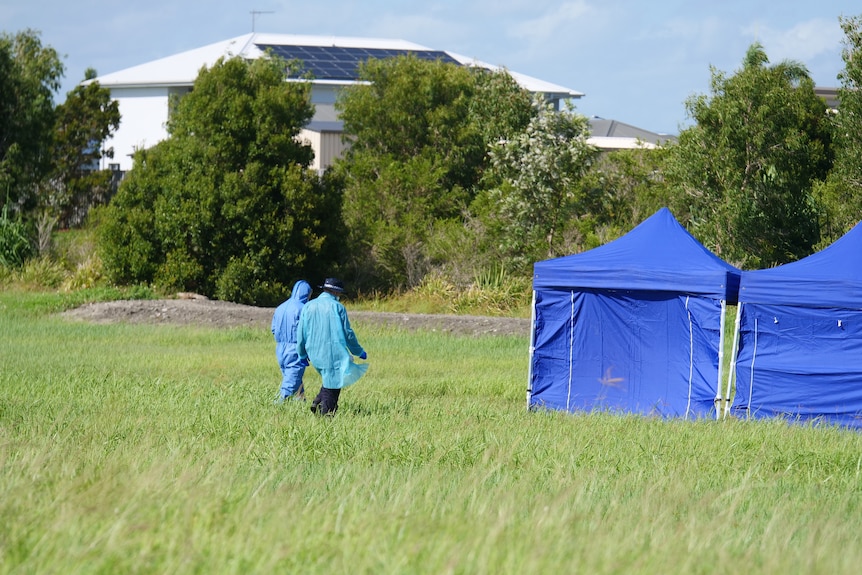 Blue tents in the middle of a grassy field, with two forensic police officers walking towards it