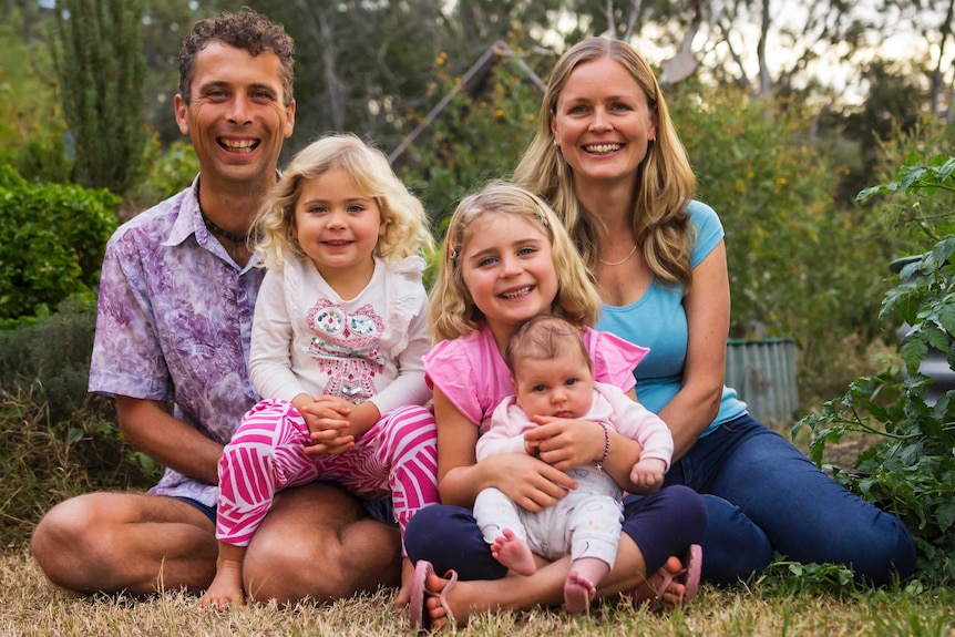 A family of five posing for a portrait in their garden with two young children and a baby, they garden and grow their own food.
