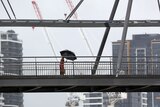 A pedestrian walks across a bridge, silhouetted against grey skies and the Brisbane skyline