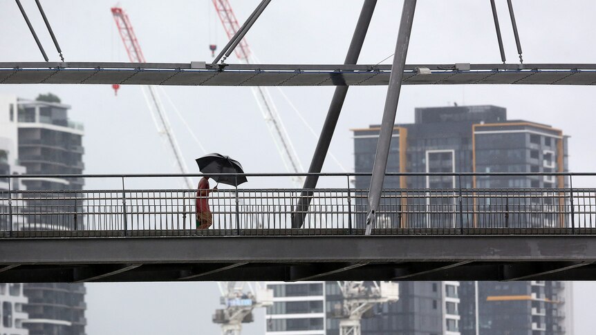 A pedestrian walks across a bridge, silhouetted against grey skies and the Brisbane skyline