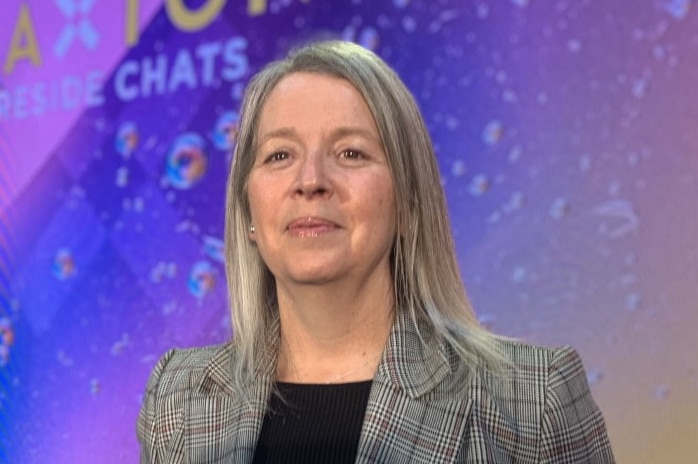 Anne Jemison smiles, standing in a grey jacket in front of a purple-coloured wall.