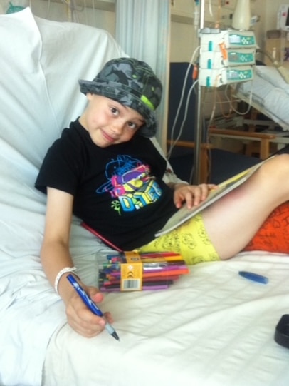 A girl wearing a hat sits on a hospital bed holding a colouring pen while posing for the camera.