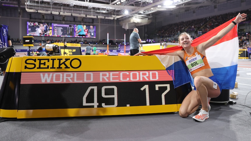 A Dutch athlete smiles and holds a national flag behind her back as she kneels next to a scoreboard with 'World Record 49.17'.