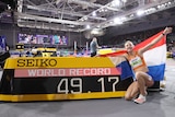 A Dutch athlete smiles and holds a national flag behind her back as she kneels next to a scoreboard with 'World Record 49.17'.