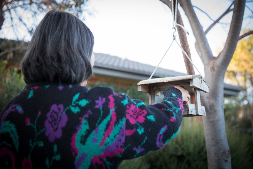 A woman with dark hair in a colourful jumper in a garden with a bird feeder. She is facing away from the camera.