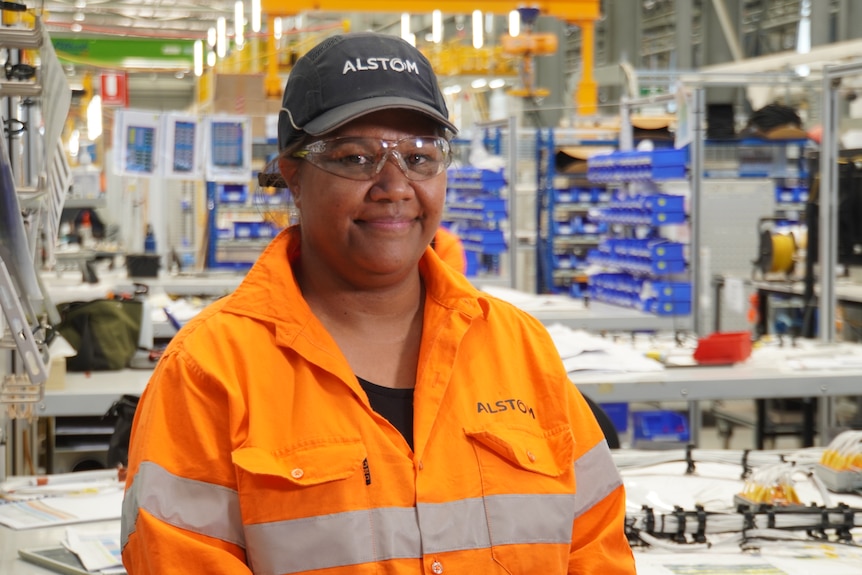 An Indigneous woman wearing an orange hi-vis shirt, safety glasses and a hat, standing in front of a cable workshop.