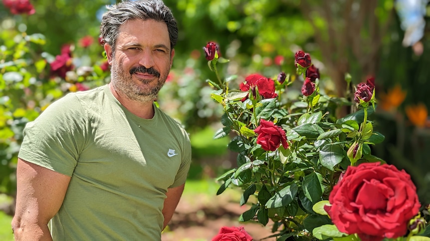Yianni Koutouzis, a Greek man, with stubble in a tight green shirt stands smiling in front of a red rose bush.