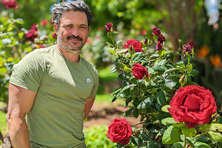 Yianni Koutouzis, a Greek man, with stubble in a tight green shirt stands smiling in front of a red rose bush.