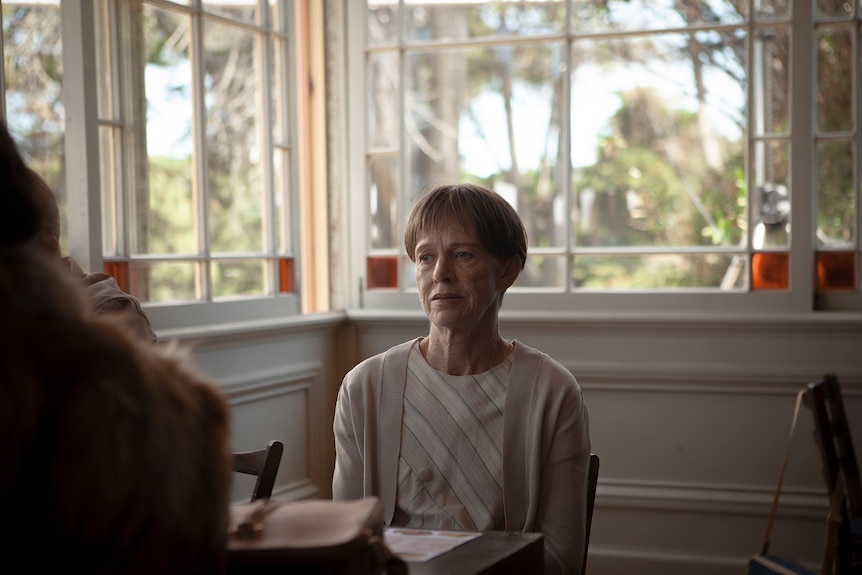 Middle-aged woman with short mousey brown hair and wearing a cream cardigan sits at a table in front of a window looking forlorn