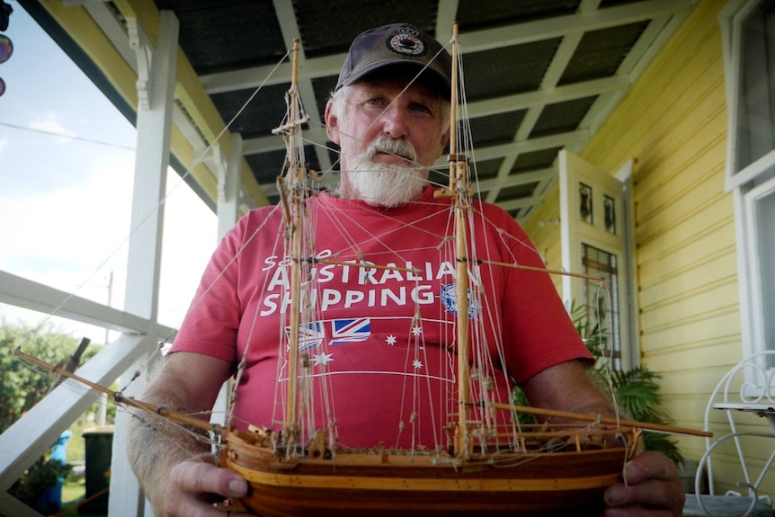 A man in a red shirt holding a model wooden ship sitting on the balcony of a house
