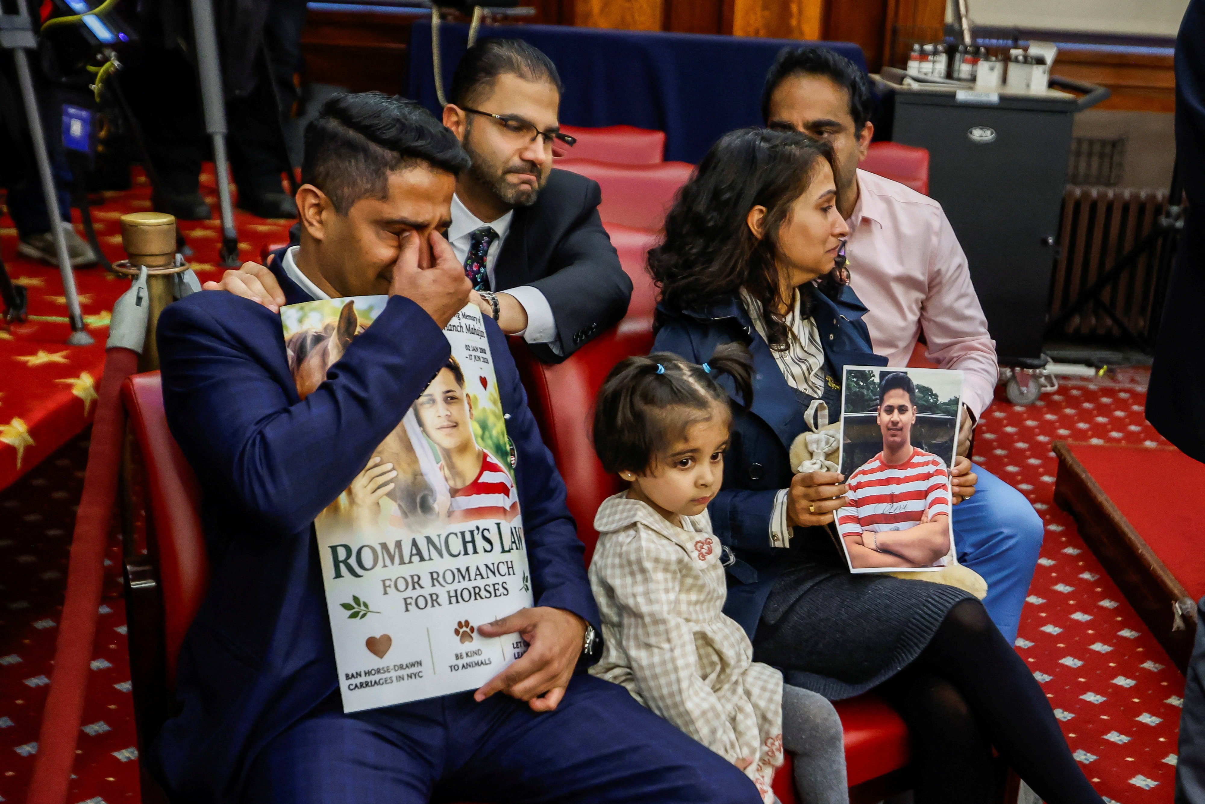 People sitting in the New York City hall, holding signs depicting Romanch Mahajan. One man is crying.