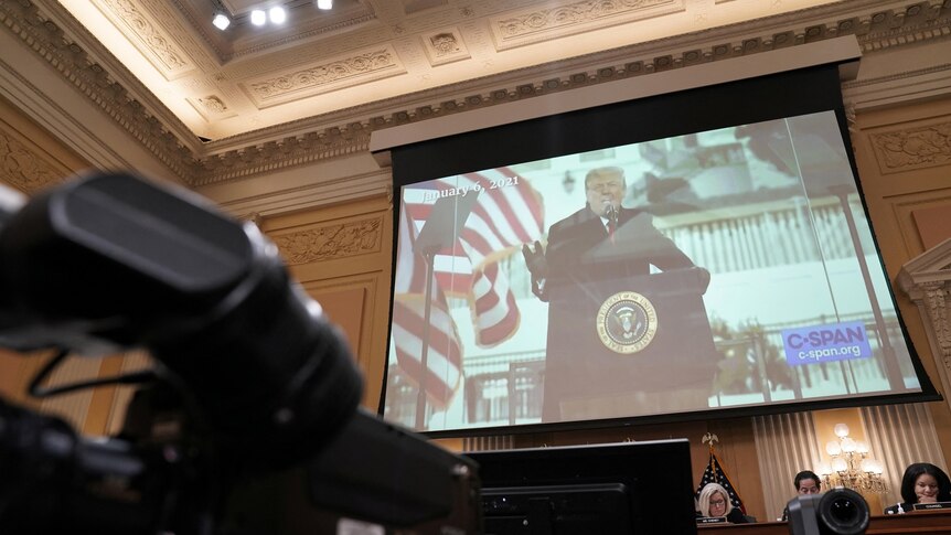 Donald Trump, speaking at a White House lectern, is seen on a screen inside an ornate room lined with cameras and people at desk