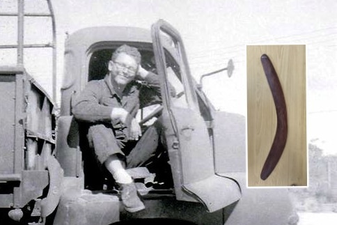 A black and white photo of a smiling soldier sitting in the open door of a truck, and a long brown boomerang.