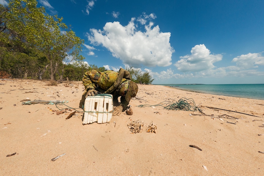 An Australian soldier leaning over and inspecting a foam float on a beach.