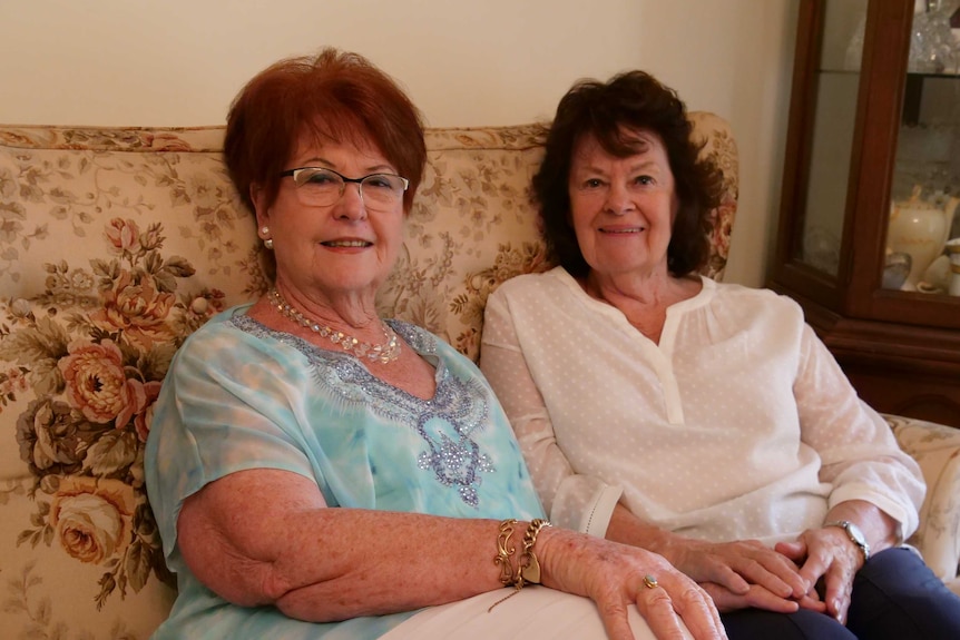 Two older women sit next to each other on a couch and smile at the camera