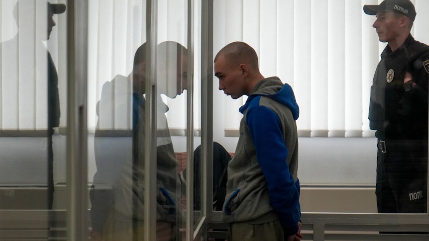 Russian Sgt. Vadim Shishimarin stands in court behind a glass wall during a hearing.
