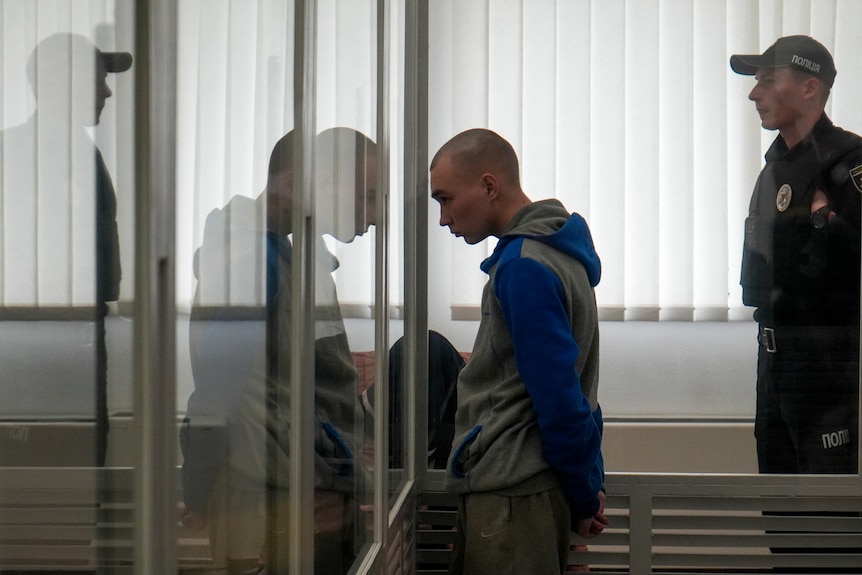 Russian Sgt. Vadim Shishimarin stands in court behind a glass wall during a hearing.