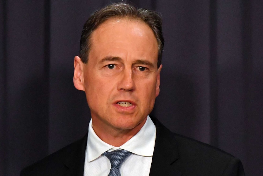 A middle aged man in a dark suit, white shirt and blue tie stands in front of a dark background.