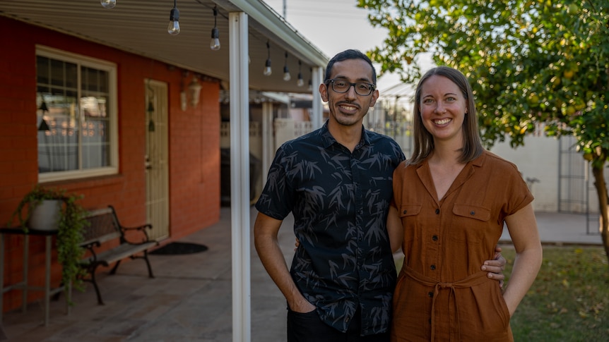 A young couple stand next to each other in a front yard. A small bench and plant stand sit against an orange wall of their house