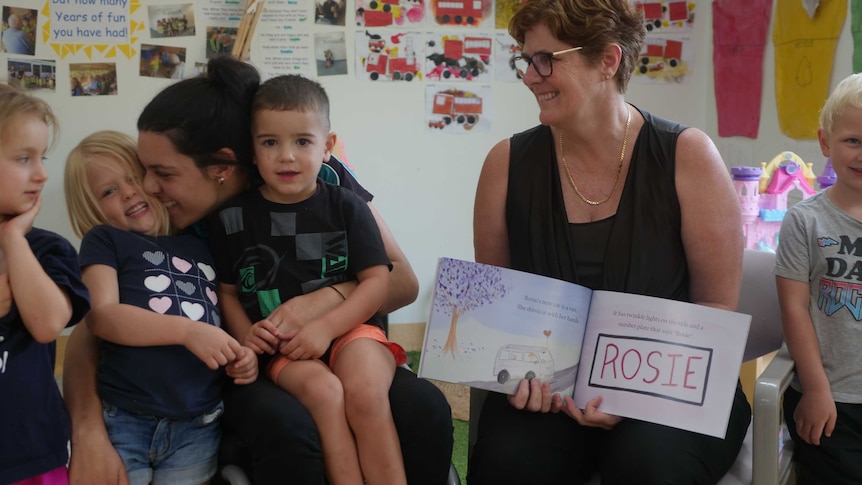 A lady in a wheelchair hugs a preschool child, while other children stand next to her smiling.