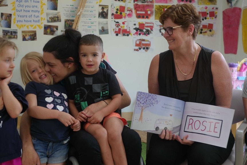 A lady in a wheelchair hugs a preschool child, while other children stand next to her smiling.