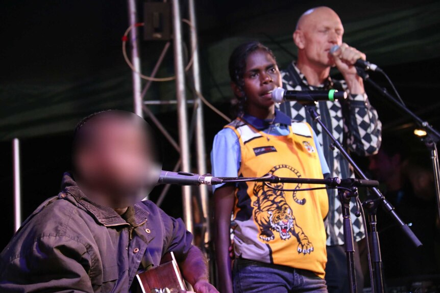 Dr G Yunupingu performs on stage sitting down with a guitar, while his daughter and Peter Garrett stand and sing.
