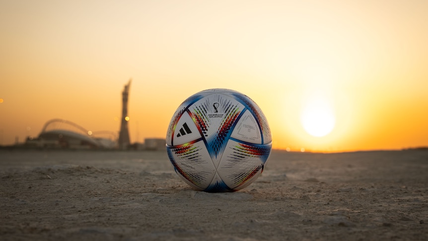 A lone football sits in the desert, in front of the setting sun and with a stadium visible in the background