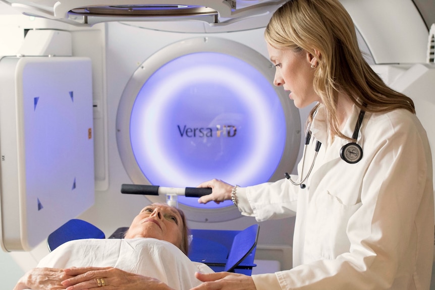 Bronwyn King wearing a white lab coat and stethoscope looks down at a patient in a radiation machine.