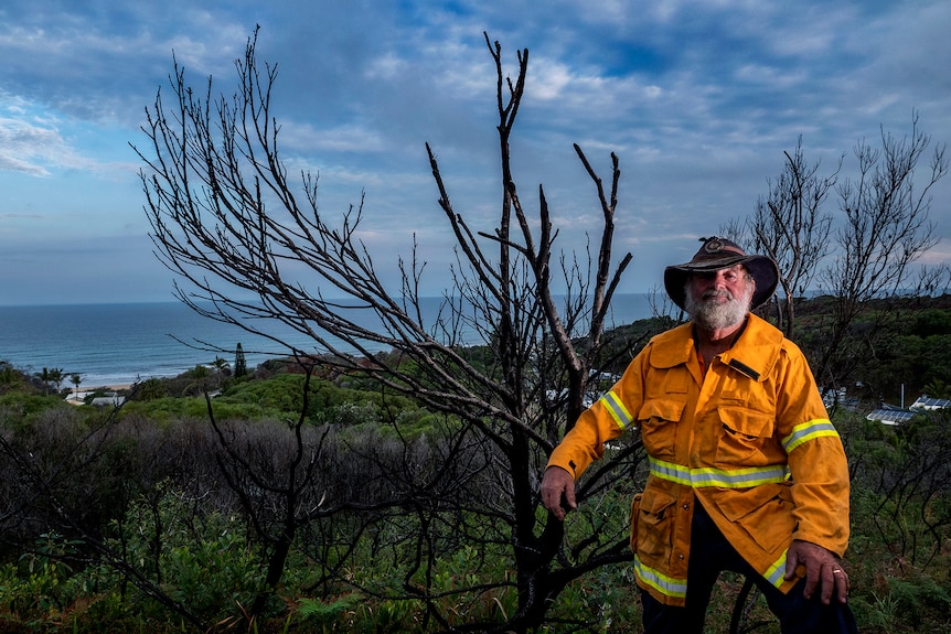man in 70s with grey beard wearing fire uniform, standing next to tree with trees and ocean in the background