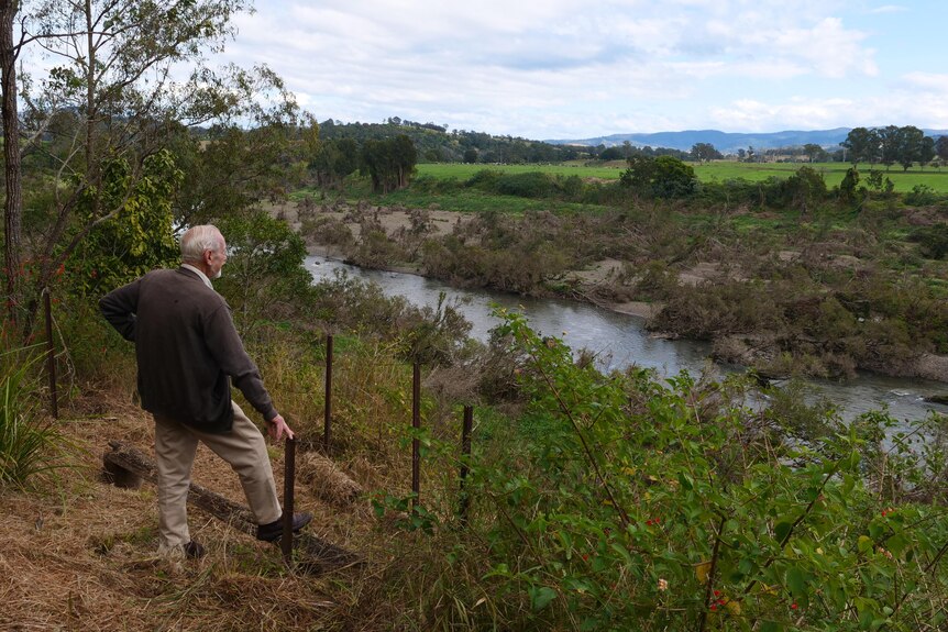 An older man stands on a rural property overlooking a river and paddocks and mountains in the distance.