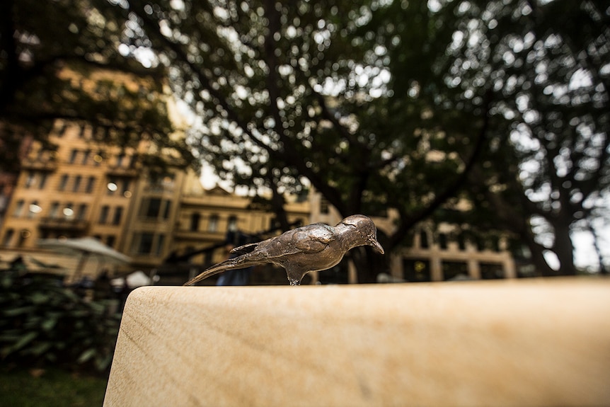 Colour photograph of a bronze bird-like sculpture standing atop of a sandstone birdbath in a Sydney CBD park.