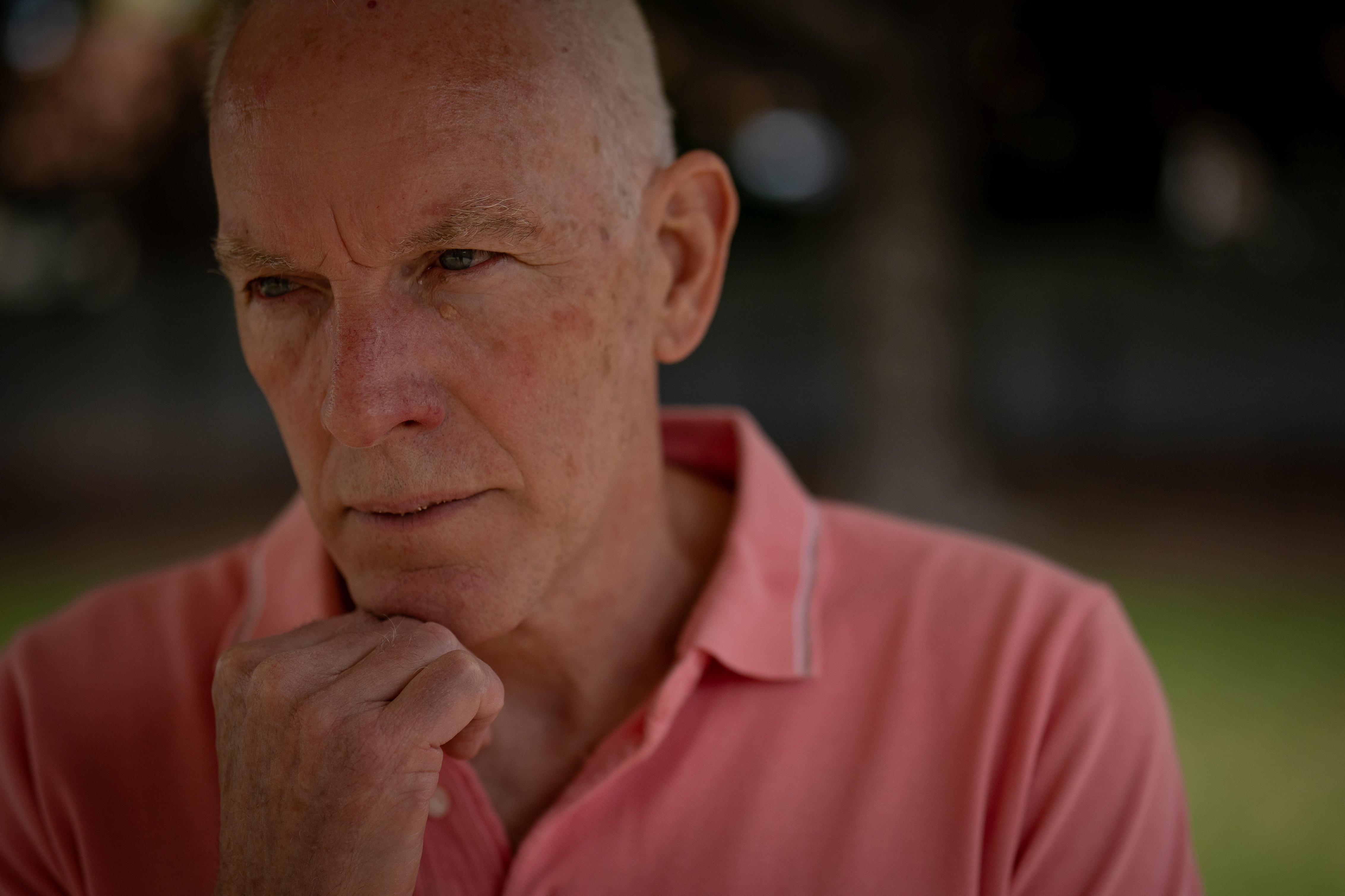 An older man wearing a salmon coloured shirt and a serious expression rests his chin in his hand.